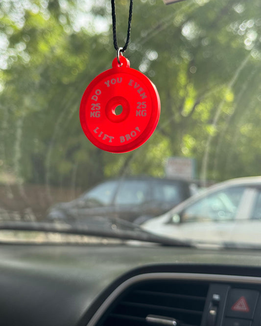 Red carabiner with text hanging from a car's rearview mirror with a blurred outdoor background.