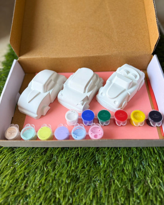 Set of white car-shaped molds with colorful liquids on a pink mat inside an open cardboard box.