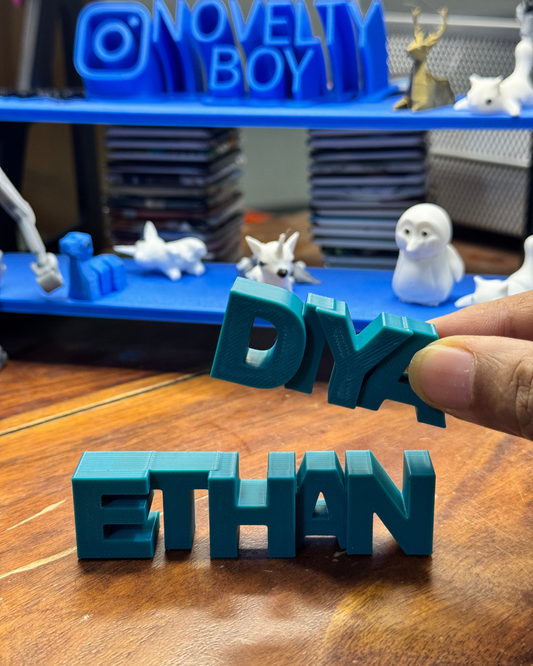 Teal 3D-printed letters spelling 'DIY ETHAN' held over a wooden surface with a blue shelf in the background.