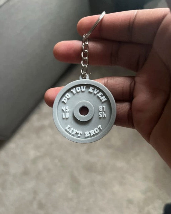 Hand holding a keychain with a humorous weightlifting disc design.