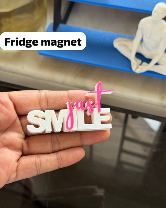 Fridge magnet shaped like 'just smile' held by a hand with a blurred background