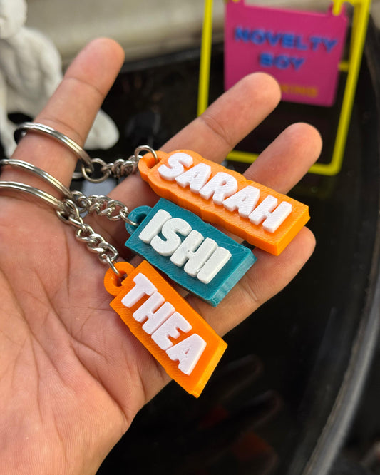 Hand holding keychains with names 'SARAH', 'ISHI', and 'THEA' against a blurred background.
