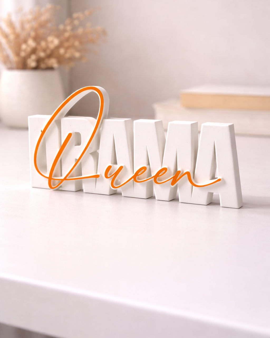 Decorative word sculpture spelling 'Queen' with white letters and orange cursive text on a white surface.