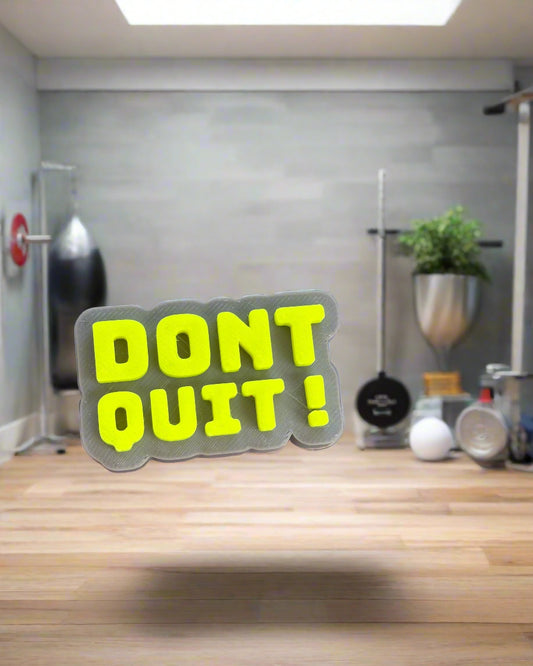 Hand holding a 'DON'T QUIT!' sticker in front of a plant pot.