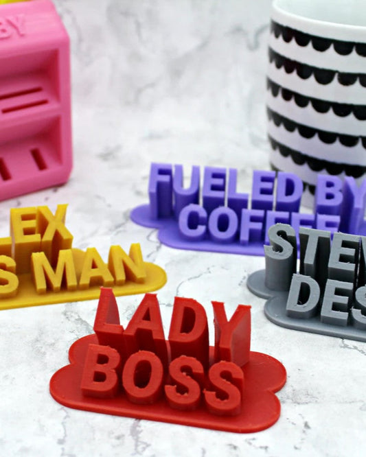 A collection of 3D printed custom desk sign boards in various colors, each with a different name and title printed on them, placed on a marble surface.