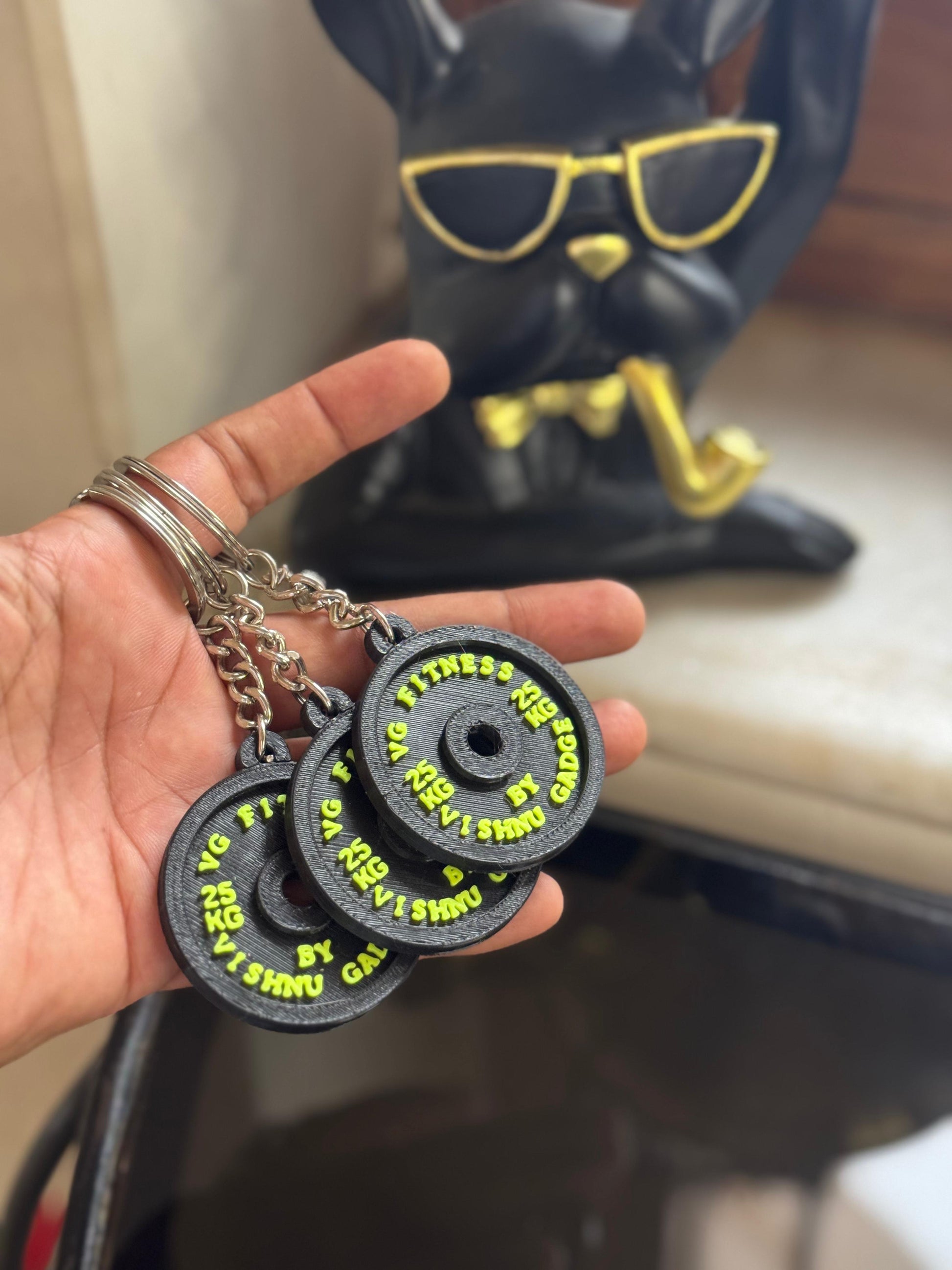Hand holding three black fitness weight plates with yellow text, with a figurine in the background.