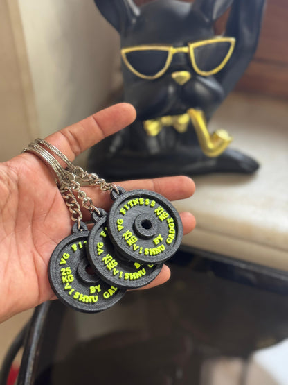 Hand holding three black fitness weight plates with yellow text, with a figurine in the background.