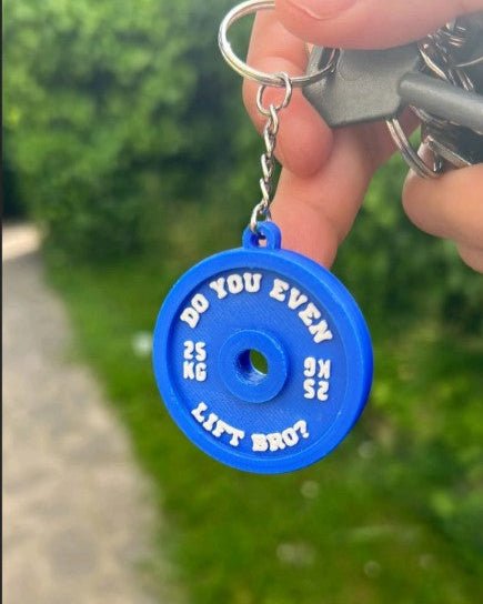 Blue keychain with a humorous weightlifting quote held by a hand outdoors.