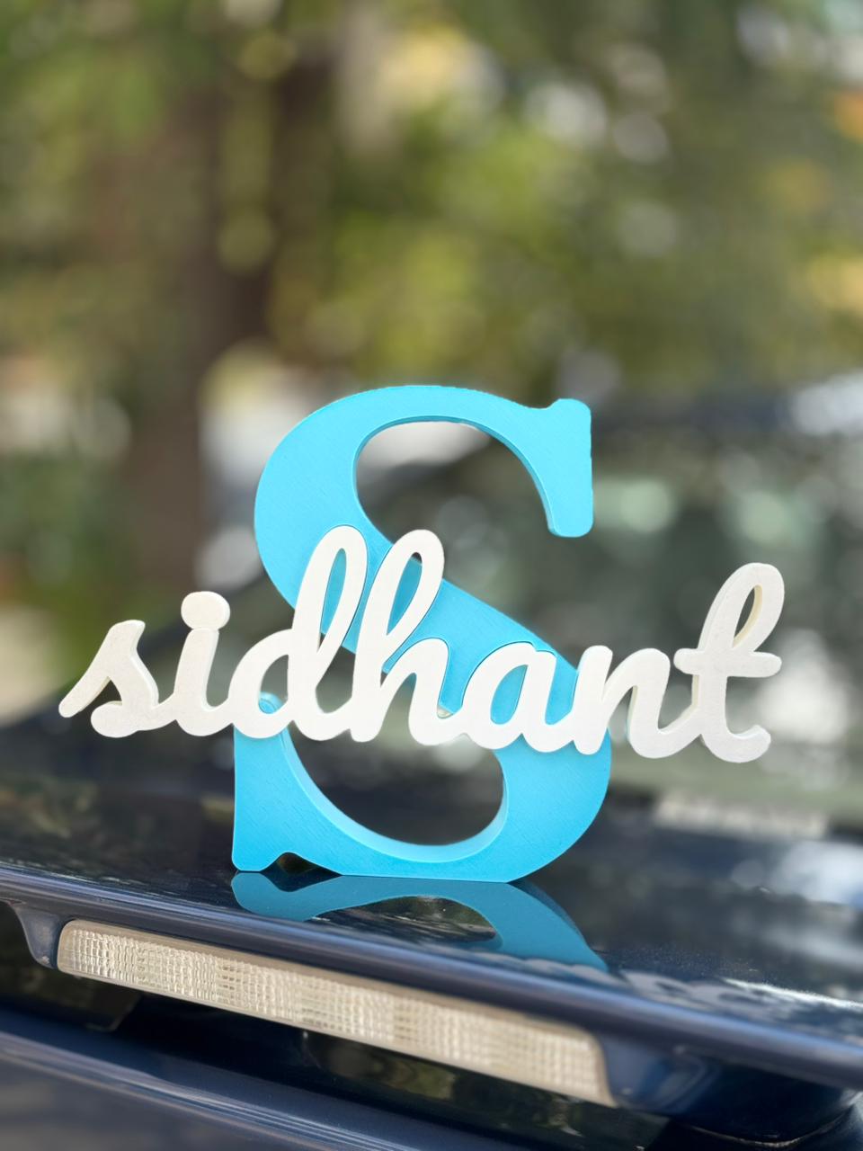 Turquoise and white decorative letter 'S' with 'sidhant' on a car window.