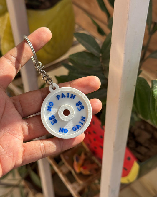 Hand holding a keychain with a white disc and blue text, surrounded by plants.