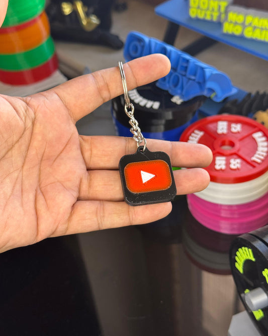 Hand holding a keychain with a play button design in front of colorful objects.